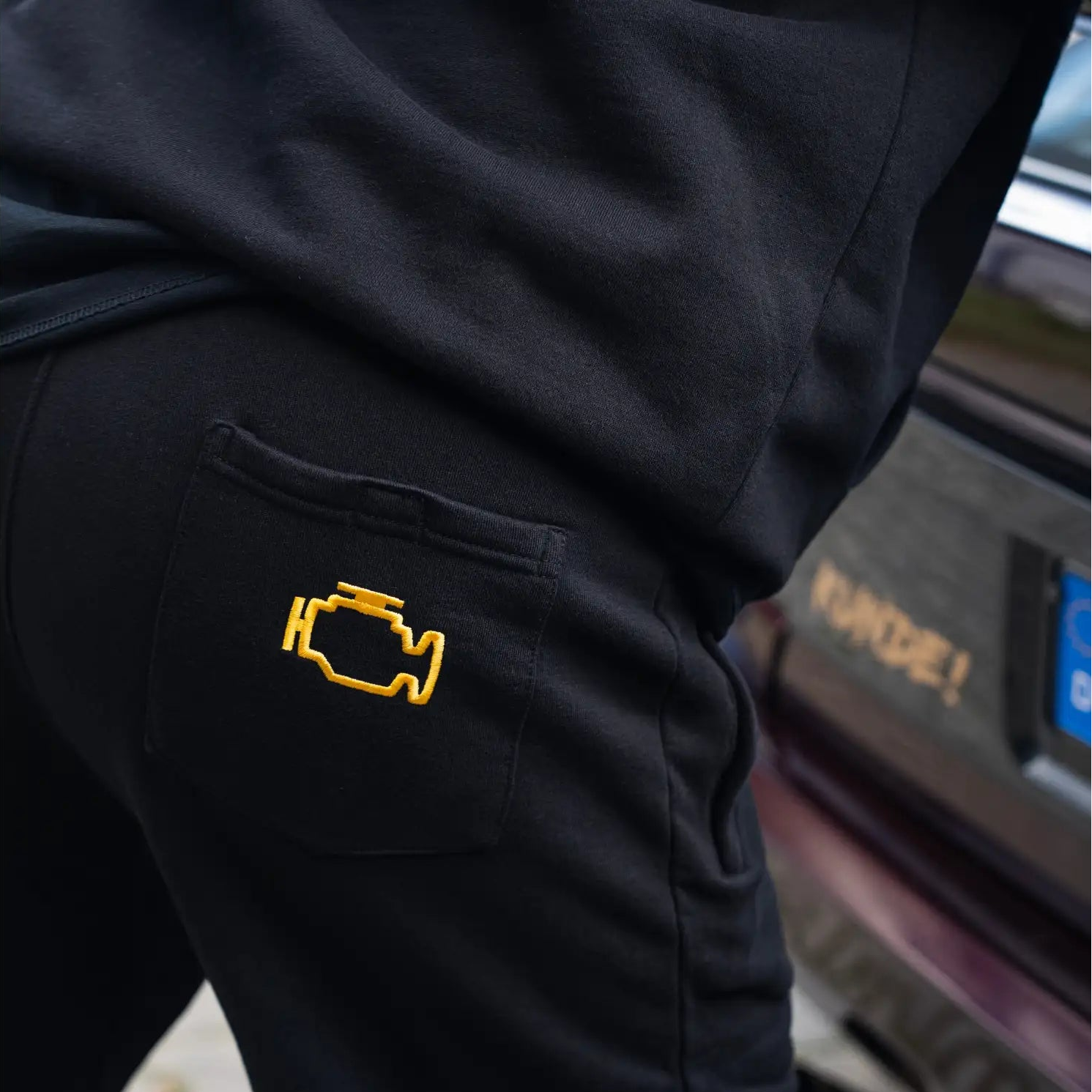 Detailaufnahme der Gesäßtasche der schwarzen Shorts mit gestickter gelber Motorkontrollleuchte.