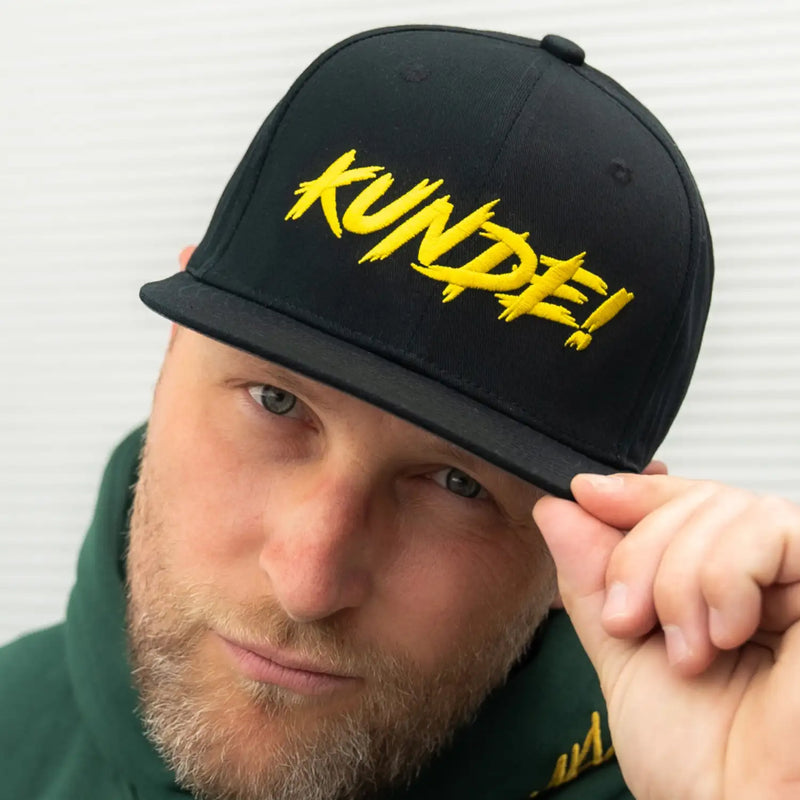 Schwarze KUNDE! Cap mit gelbem Stick auf der Vorderseite, von vorn am Model getragen.
