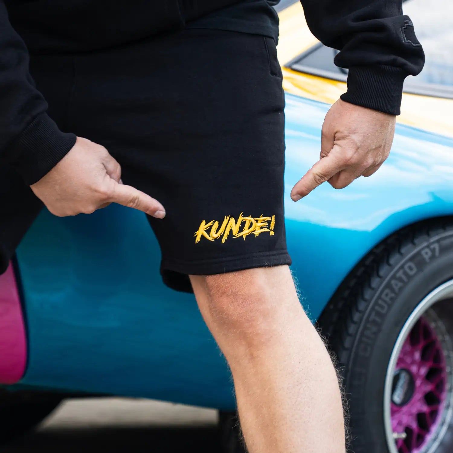 Schwarze „KUNDE!“ Shorts mit gelbem Stick vorn am Bein, am Model getragen.